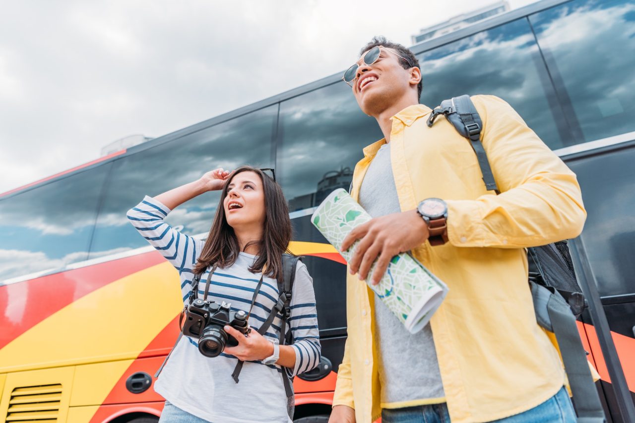 Woman holding digital camera and looking away with mixed race man holding map near travel bus.jpg